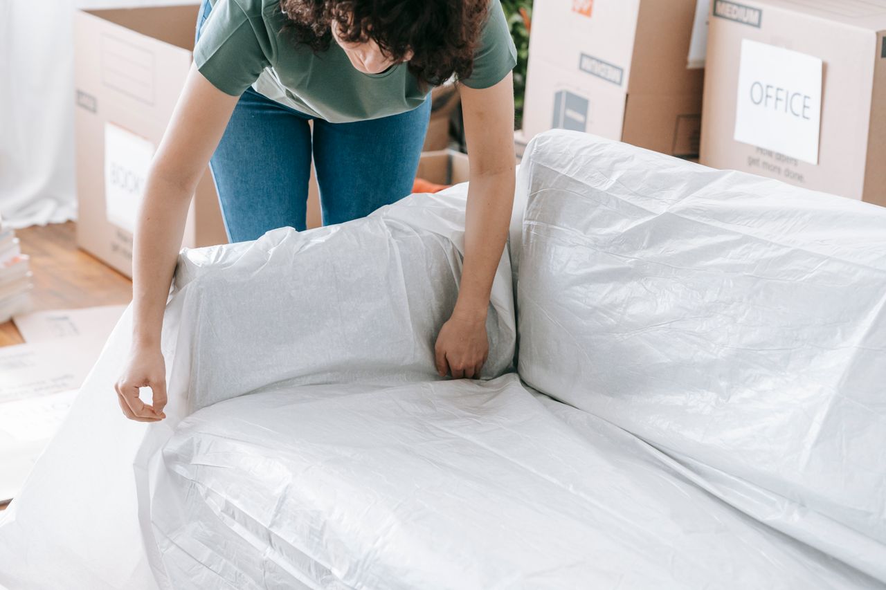 about-01 A woman carefully wraps a sofa in protective material while packing boxes in a bright room.