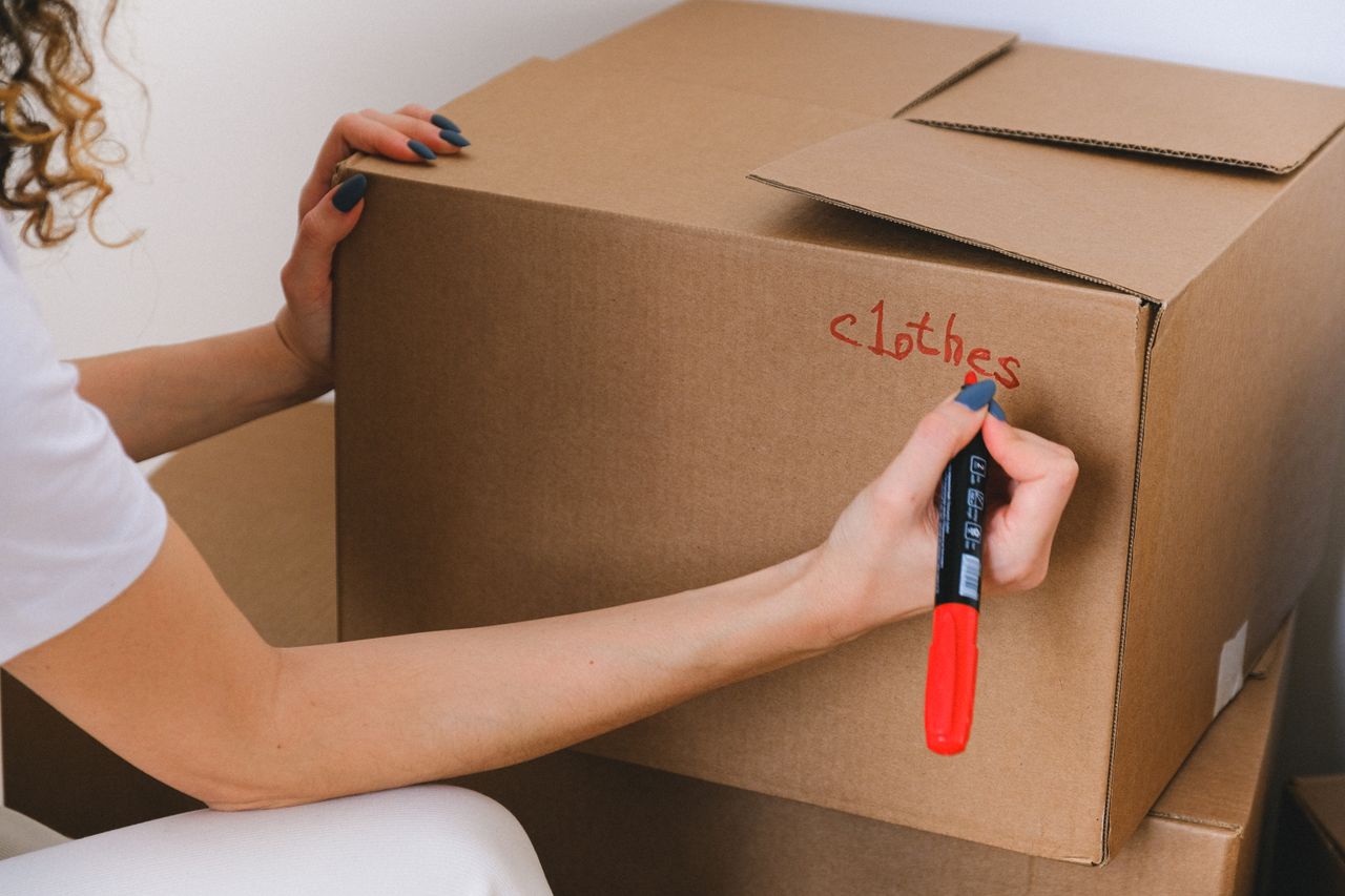hero-02 Close-up of a woman labeling a cardboard box with a red marker indoors.