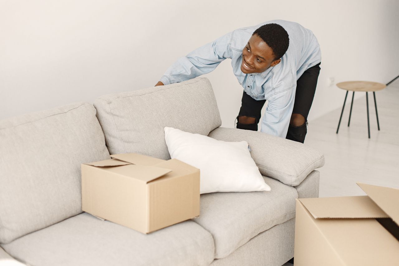 hero-01 Young man smiling while arranging cardboard boxes in a modern living room.